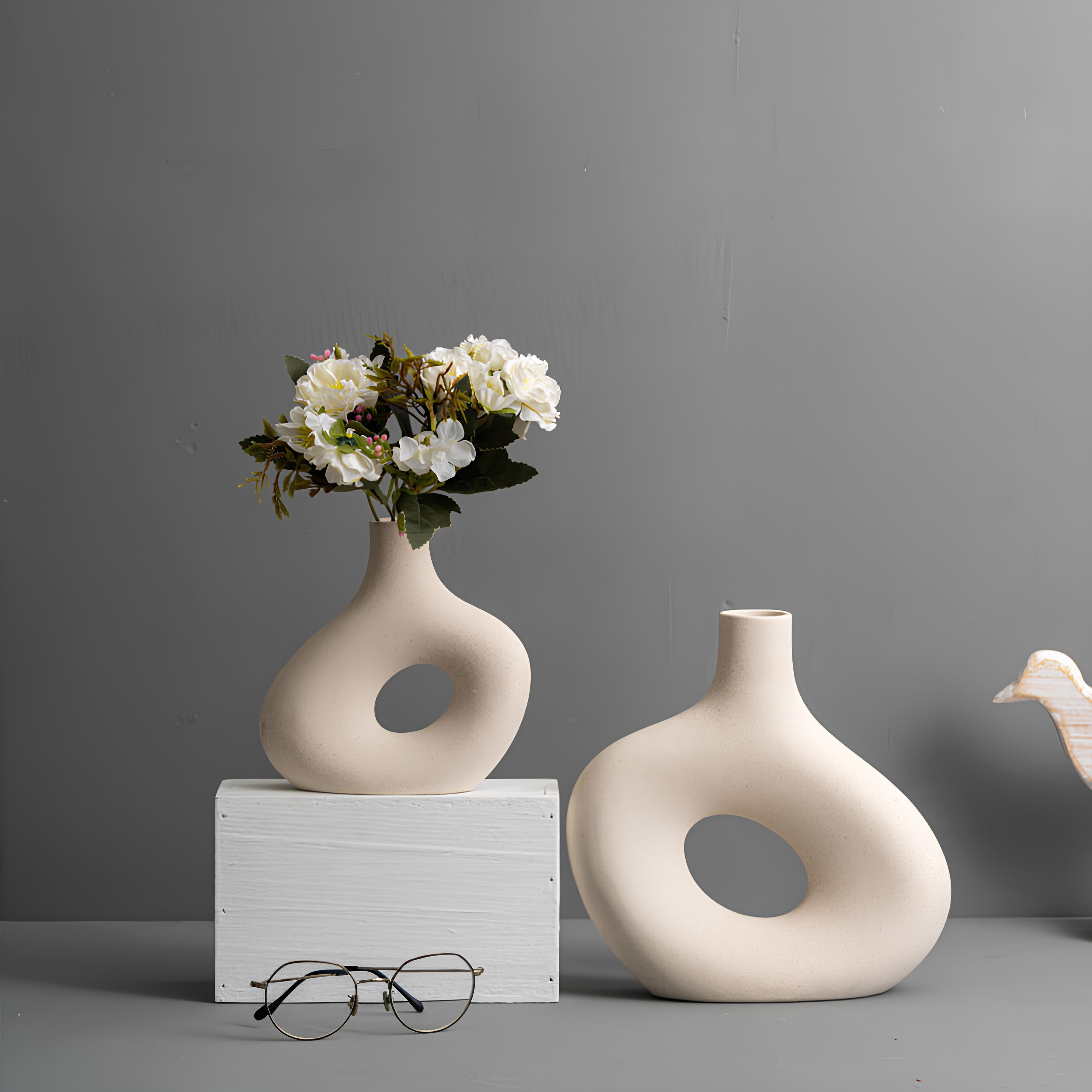 Two beige vases with circular designs, one holding flowers, on a gray surface with a neutral background.